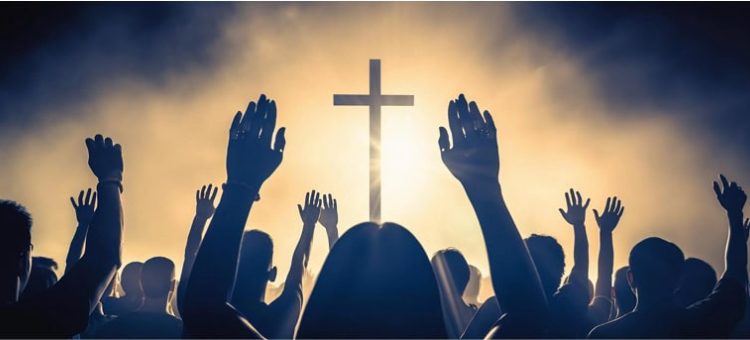 People raising hands in worship with a cross in the background at sunset.