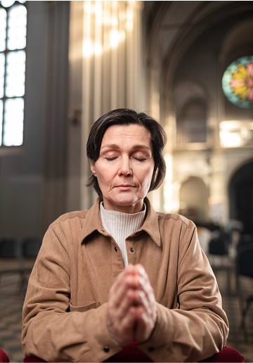 A woman praying with her eyes closed inside a church, hands clasped in prayer.
