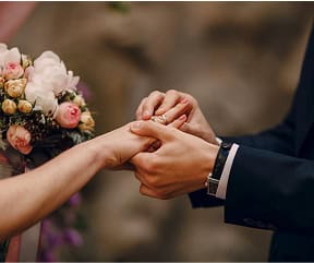 A couple holding hands during a wedding ceremony with a bouquet of flowers in the background.