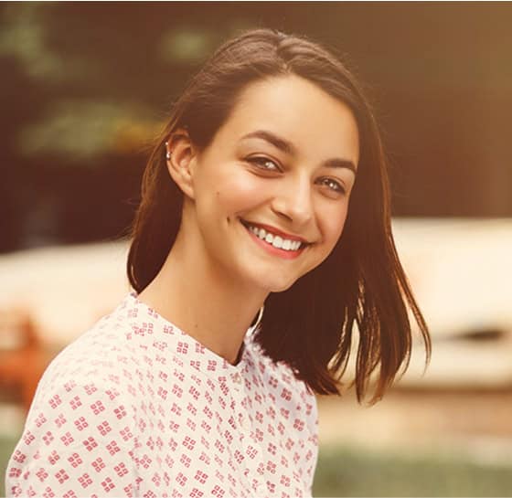 A woman smiling outdoors with natural background, wearing a white and pink patterned top.