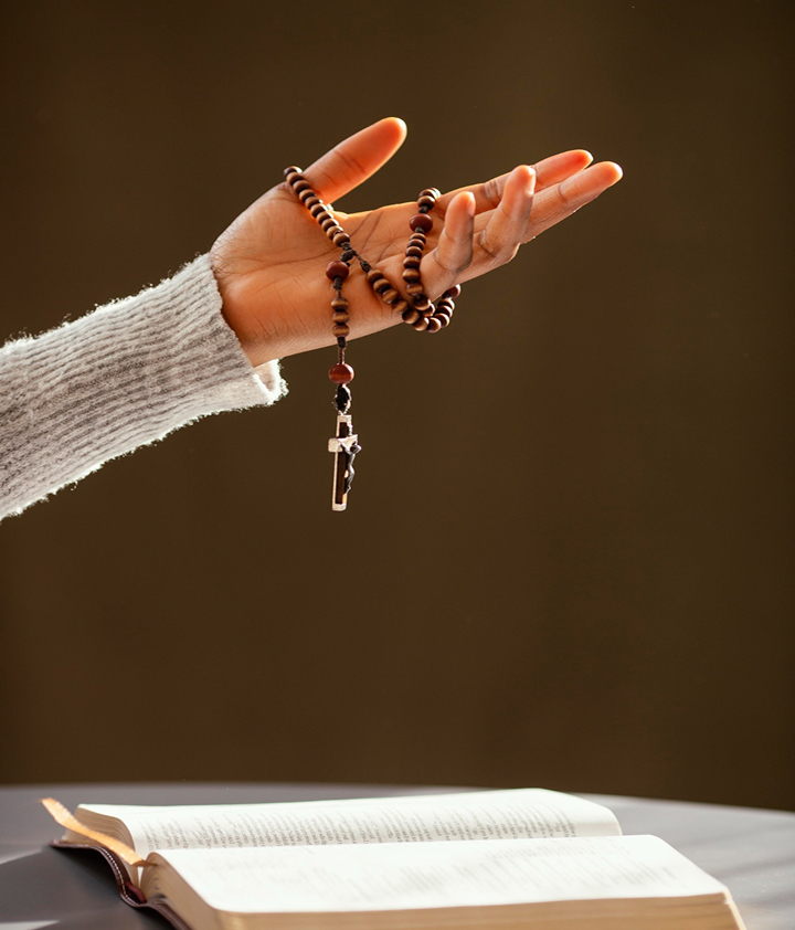 Hand holding wooden prayer beads with cross, over open book, warm background.