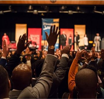 A vibrant conference with attendees raising hands on stage.